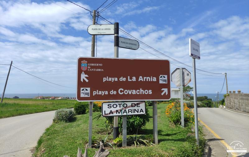Cruce y camino de acceso señalizado hacia la playa de Covachos desde la CA-231, con el prado de aparcamiento al fondo