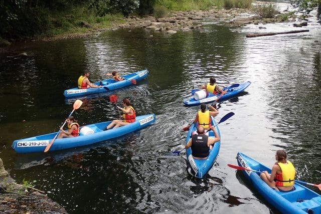 Group of canoeists paddling in a line through a stretch with a small gentle riffle, helmets and life jackets on, summer midday.
